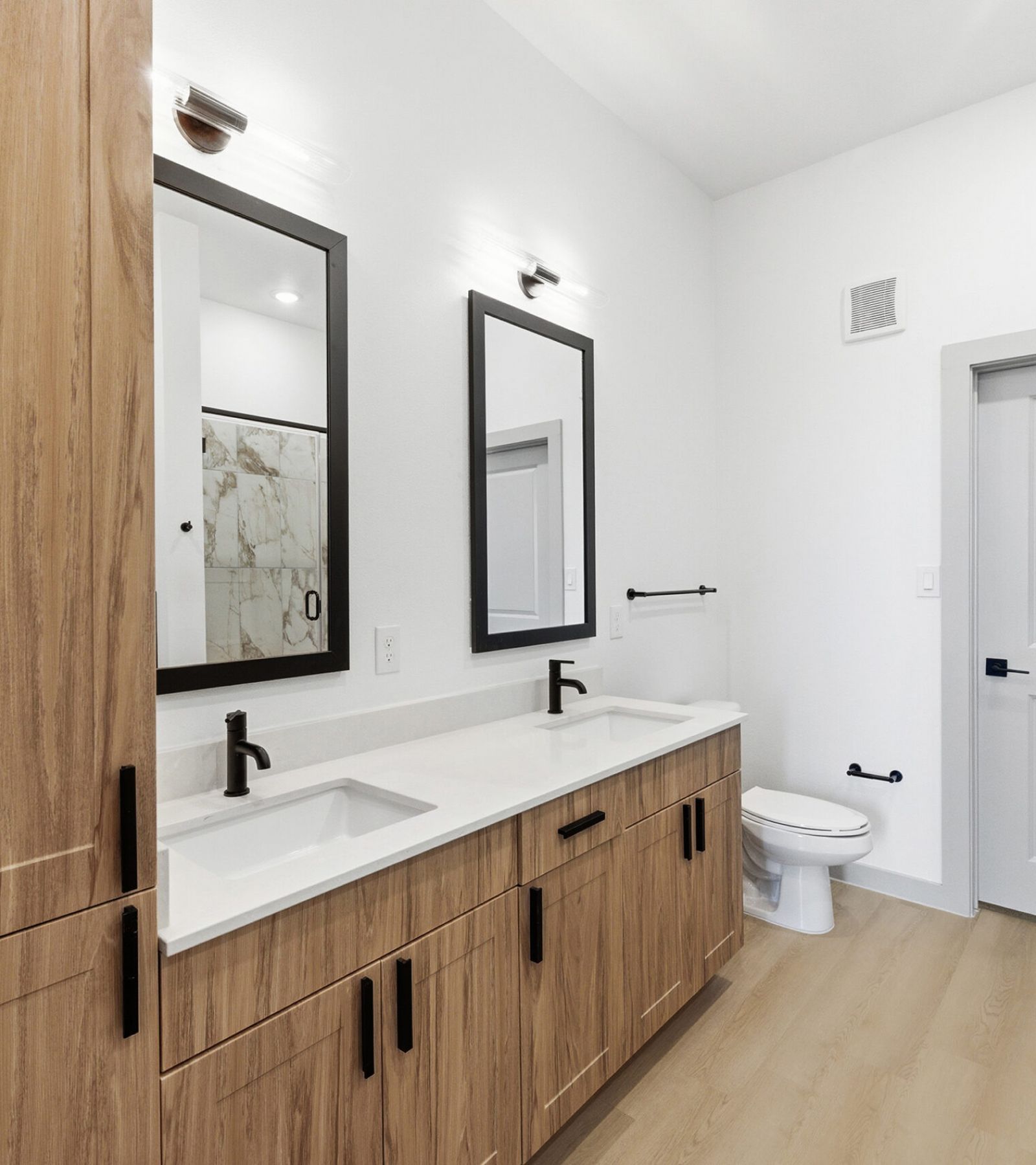 Wixby House Modern bathroom with wood cabinets, double sink vanity, black fixtures, and a toilet next to a gray door.